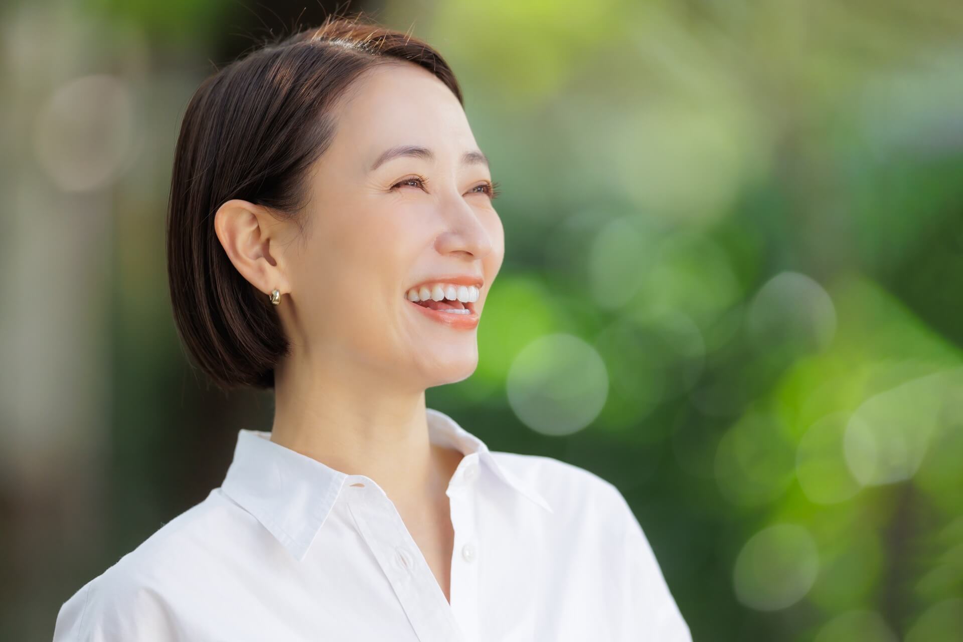白いシャツを着て新緑の中で明るくほほえむショートヘアの女性