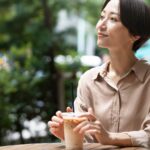 カフェのテラス席でアイスコーヒーを手に穏やかにほほえむベージュのシャツの女性