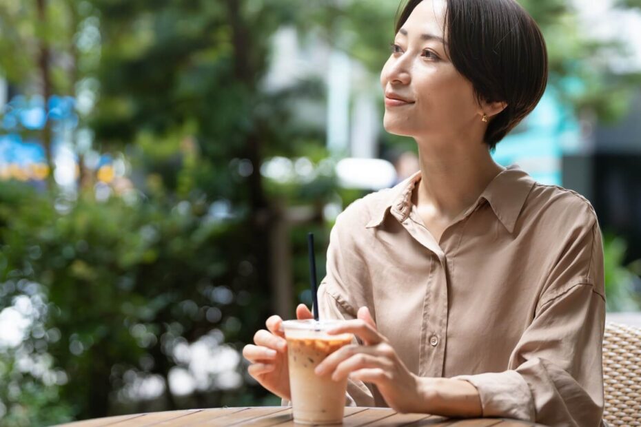 カフェのテラス席でアイスコーヒーを手に穏やかにほほえむベージュのシャツの女性