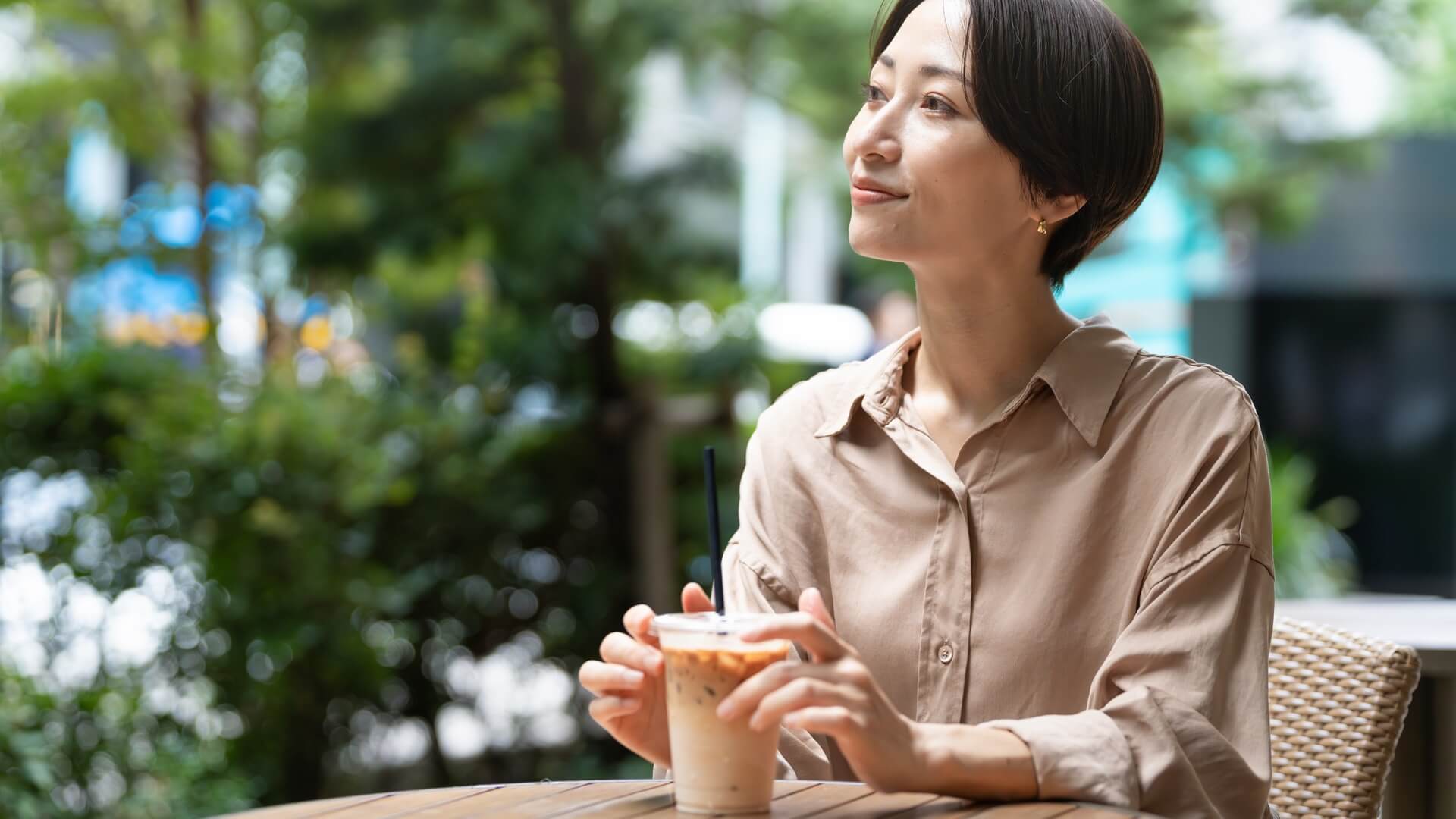 カフェのテラス席でアイスコーヒーを手に穏やかにほほえむベージュのシャツの女性