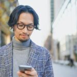 街中でスマートフォンを見ながら待ち合わせをするジャケット姿の男性