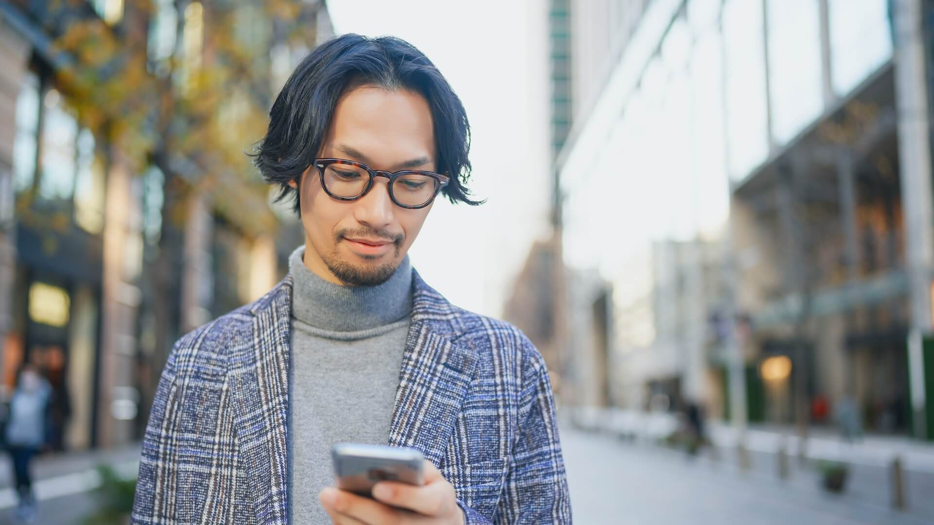街中でスマートフォンを見ながら待ち合わせをするジャケット姿の男性