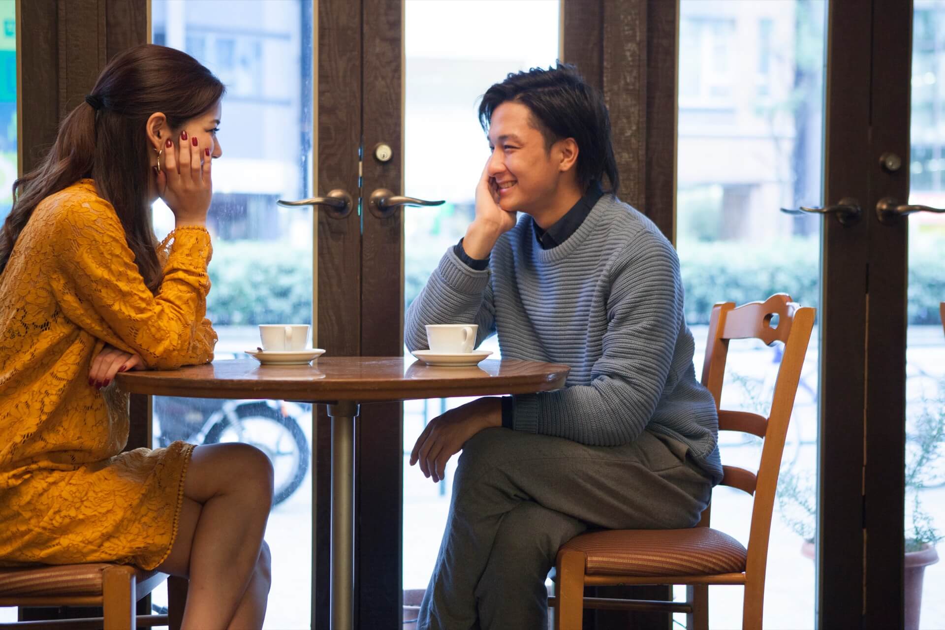 カフェのテーブルを挟んで向かい合い、会話する男女。男性はグレーのニットを着ている