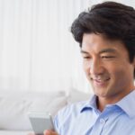 水色のシャツを着てソファでスマートフォンを見ながらほほえむ男性