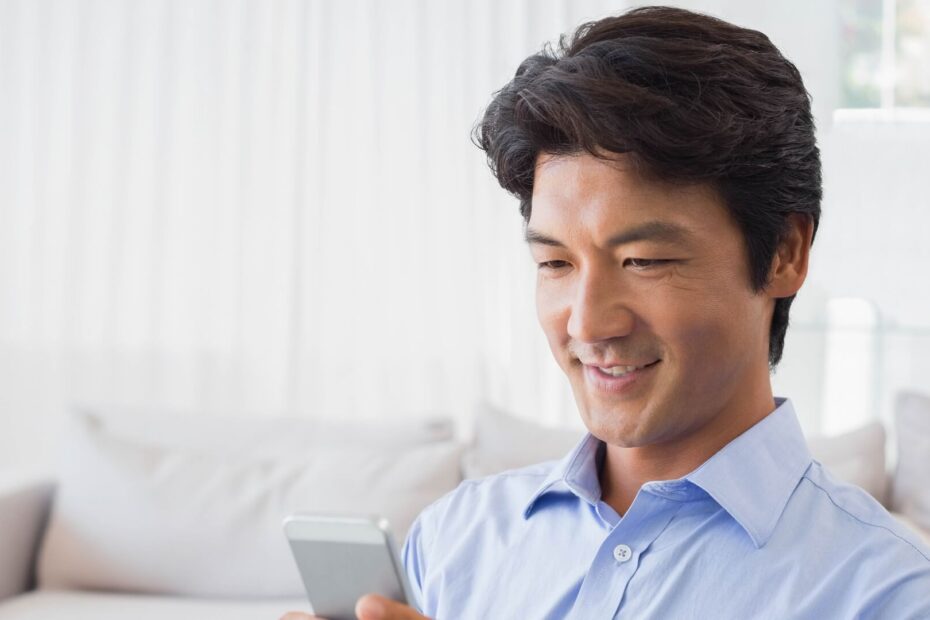 水色のシャツを着てソファでスマートフォンを見ながらほほえむ男性
