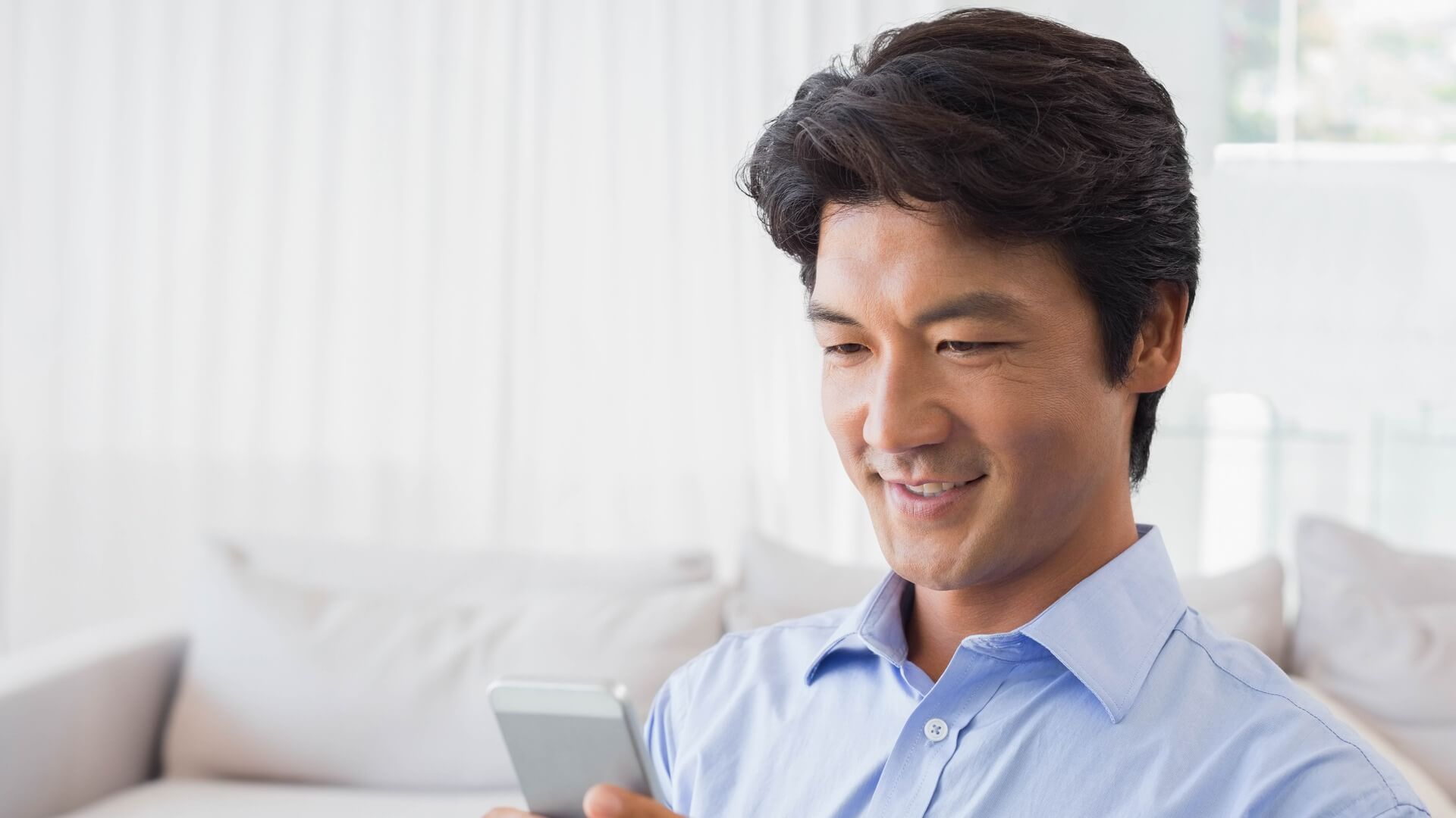 水色のシャツを着てソファでスマートフォンを見ながらほほえむ男性