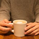 グレーのニットを着た男性が、カフェのテーブルでカップを両手で持っている様子