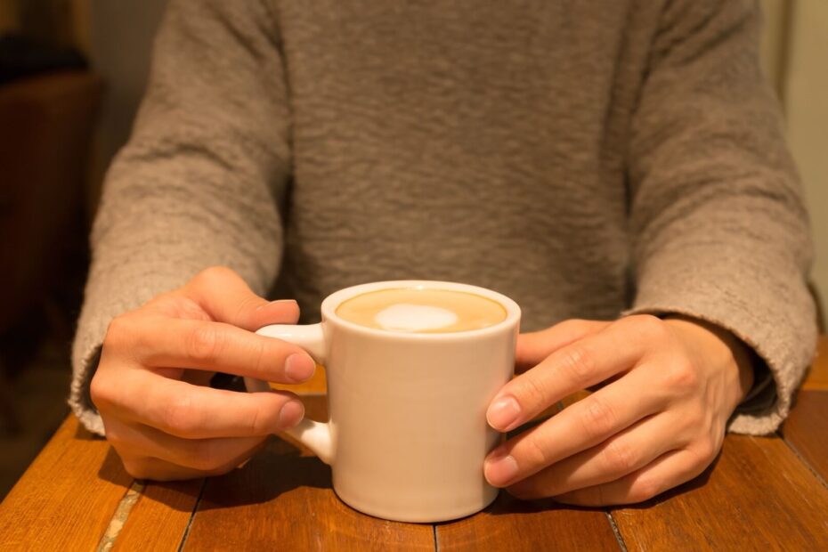グレーのニットを着た男性が、カフェのテーブルでカップを両手で持っている様子