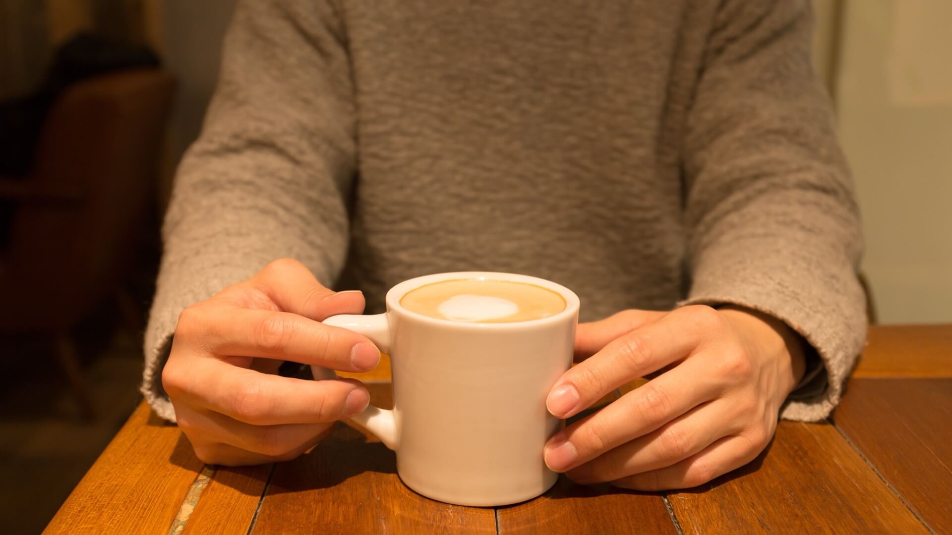 グレーのニットを着た男性が、カフェのテーブルでカップを両手で持っている様子