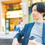 ネイビーのジャケットにグレーのニットを合わせ、街のベンチでコーヒーを持つ男性
