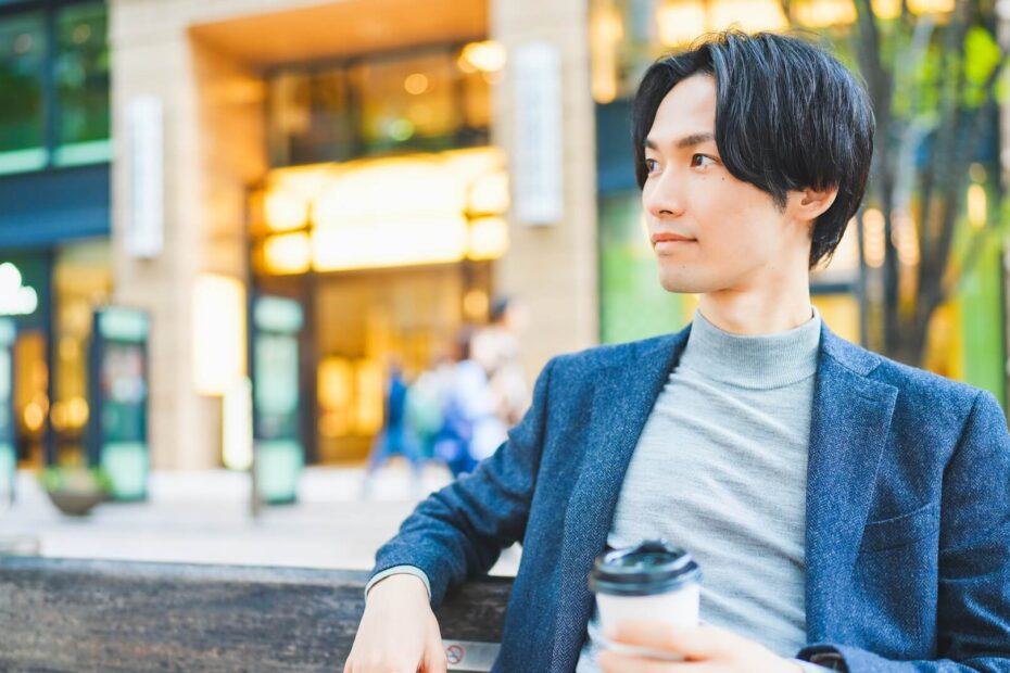ネイビーのジャケットにグレーのニットを合わせ、街のベンチでコーヒーを持つ男性