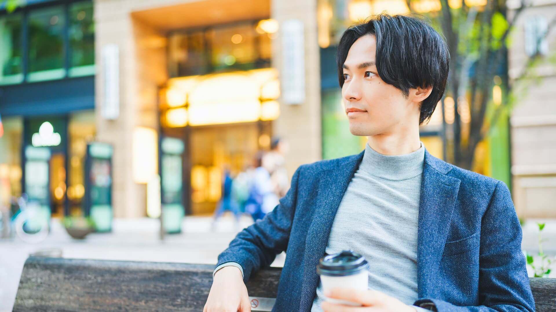 ネイビーのジャケットにグレーのニットを合わせ、街のベンチでコーヒーを持つ男性
