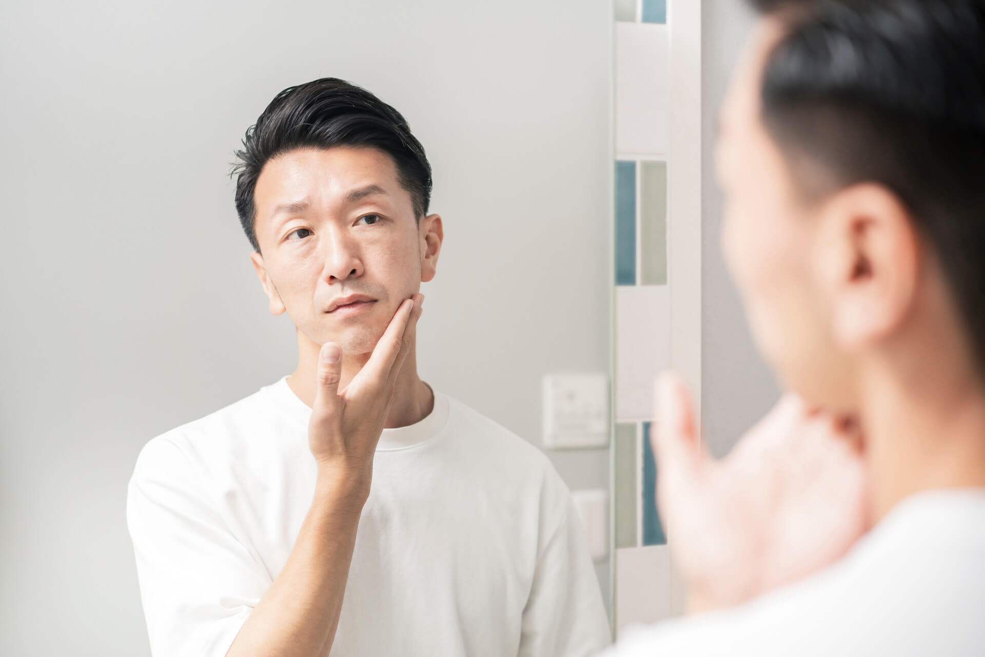 洗面所の鏡を見ながらあごに手を当てて肌の状態を確かめる男性