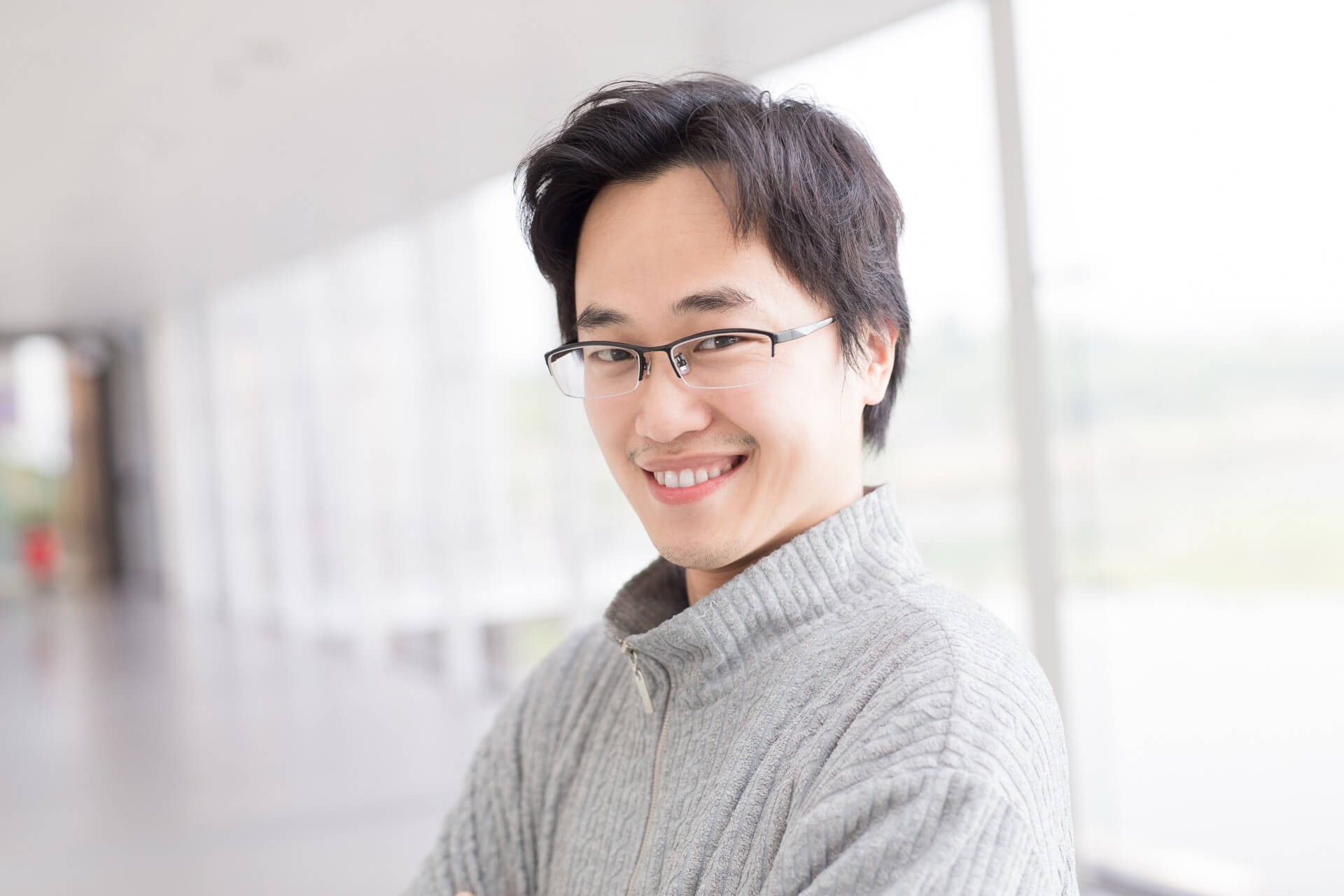 グレーのハーフジップニットを着て、明るい室内で微笑む眼鏡の男性