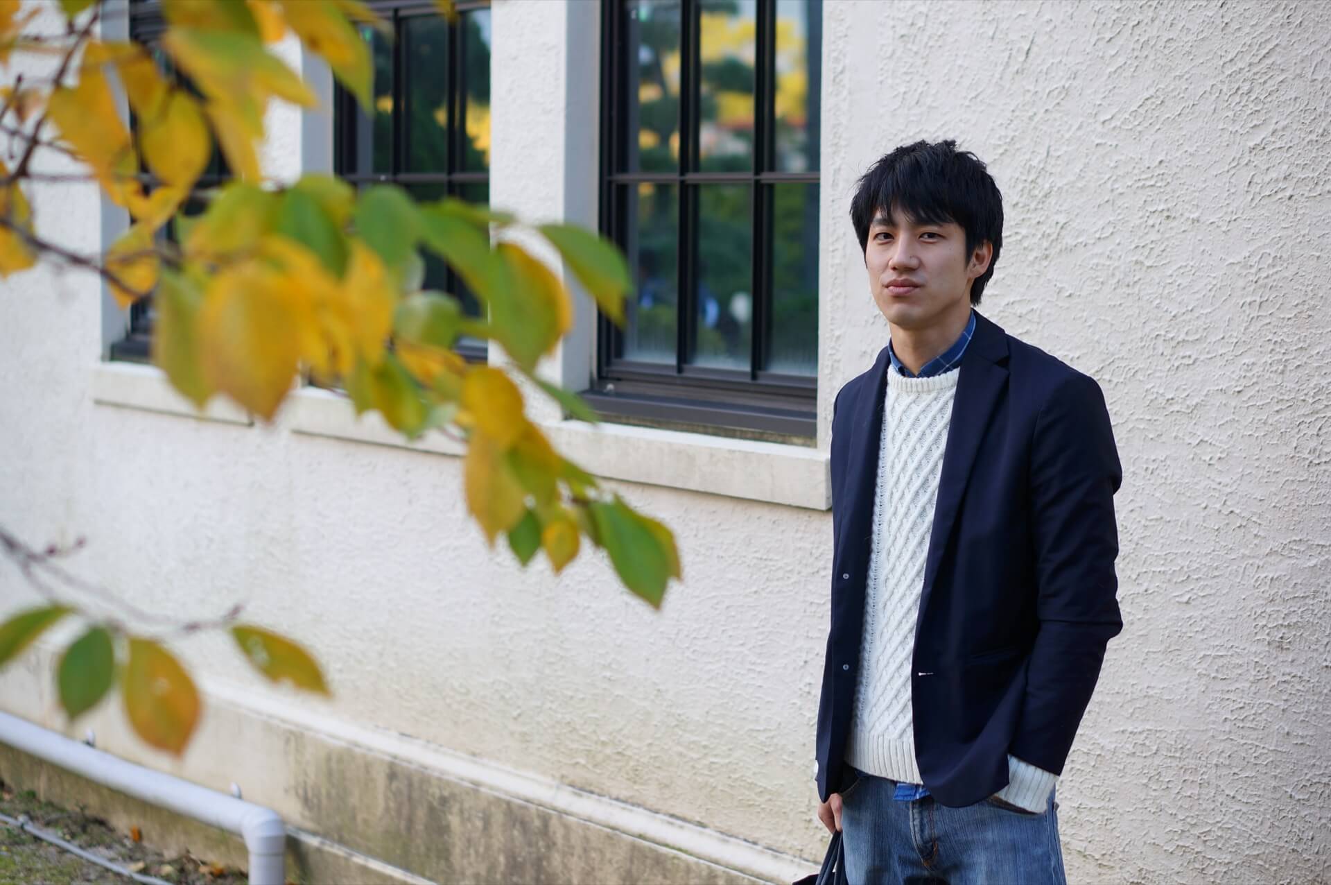 明るい色のニットにネイビーのジャケットを羽織り、屋外に立つ男性