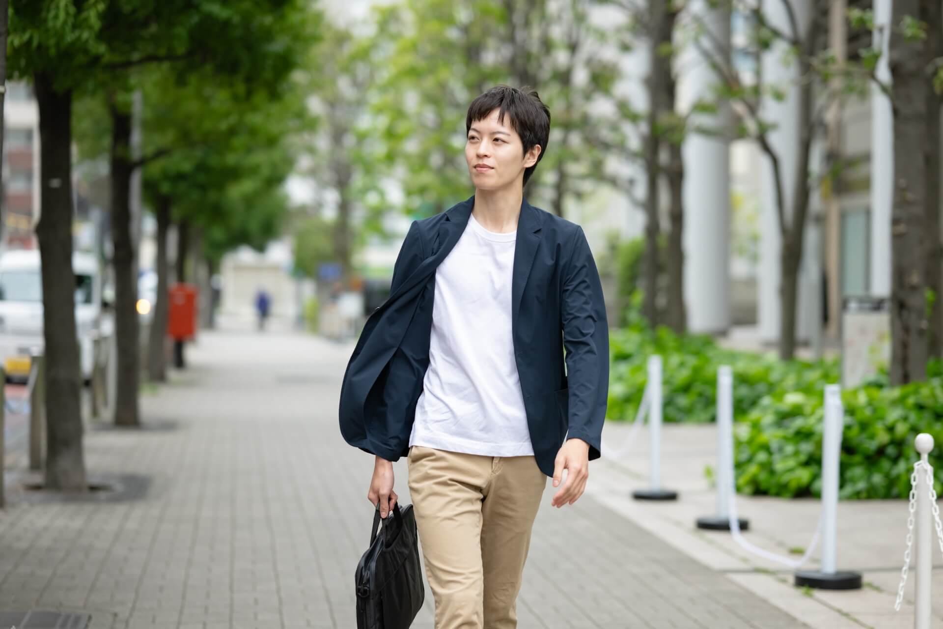 ネイビーのジャケットに白いTシャツを合わせて並木道を歩く男性