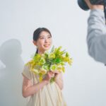 白い壁の前で花束を持ち、カメラマンにプロフィール写真を撮影してもらう女性