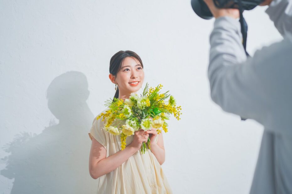 白い壁の前で花束を持ち、カメラマンにプロフィール写真を撮影してもらう女性