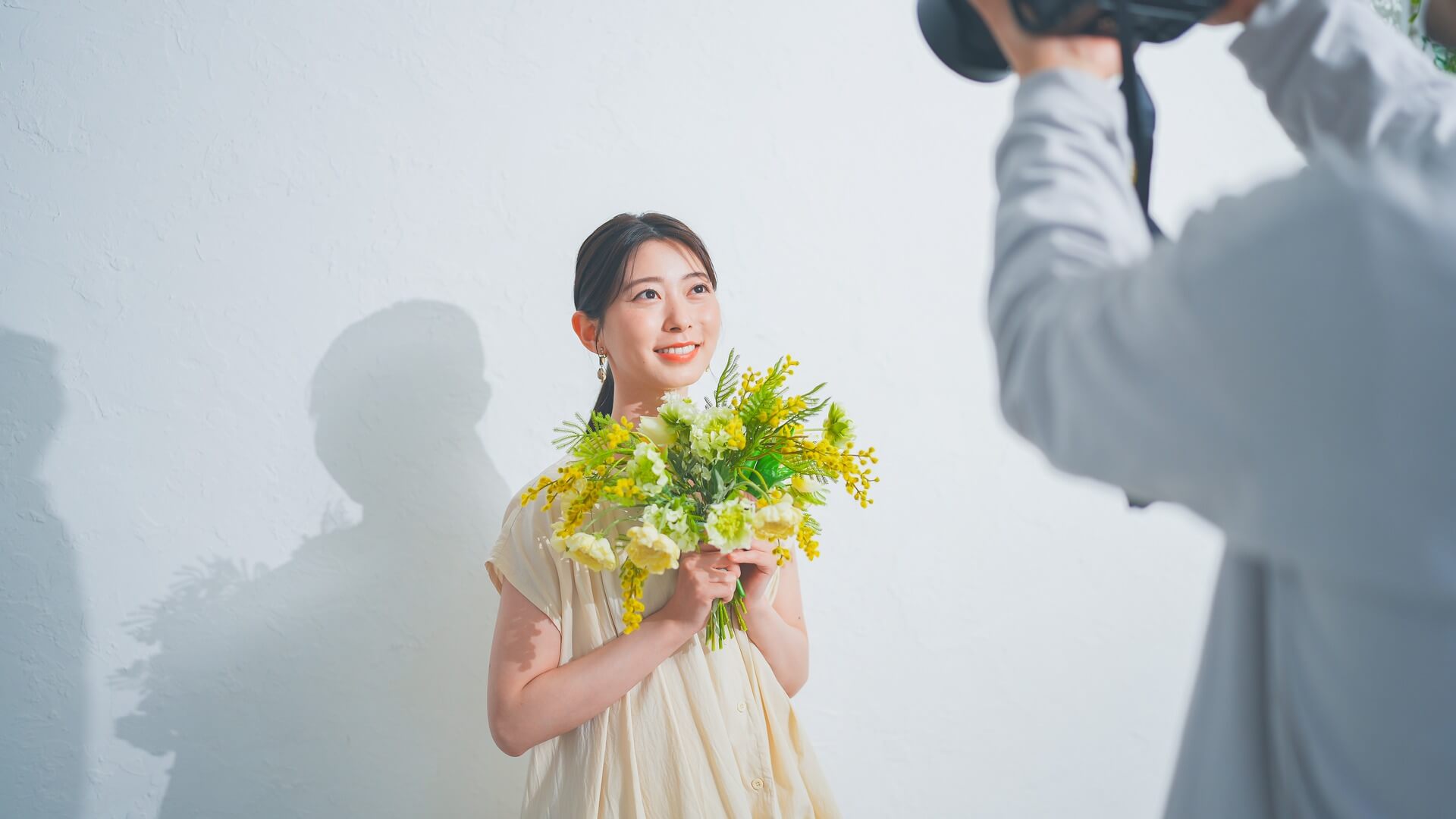 白い壁の前で花束を持ち、カメラマンにプロフィール写真を撮影してもらう女性