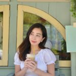 カフェの前で白いワンピースを着てアイスドリンクを手にほほえむ女性
