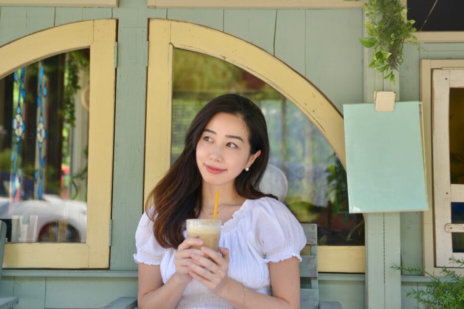 カフェの前で白いワンピースを着てアイスドリンクを手にほほえむ女性