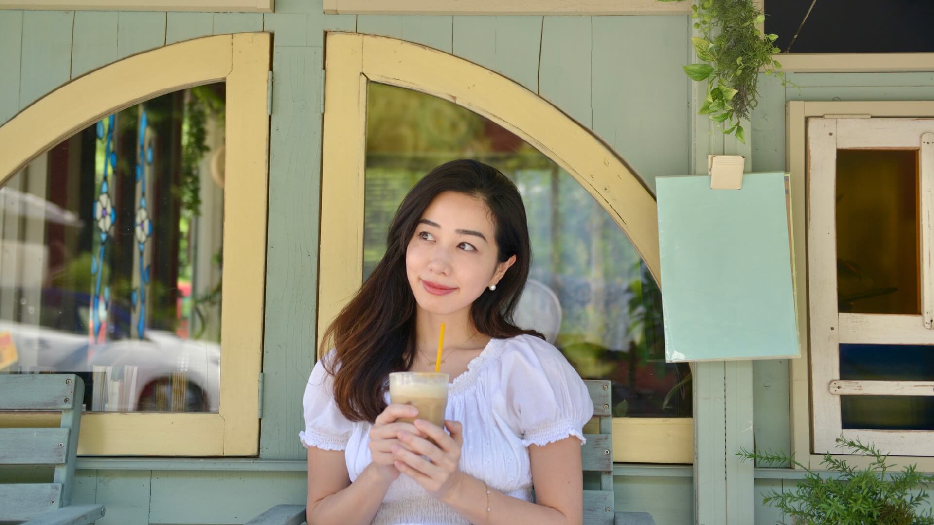 カフェの前で白いワンピースを着てアイスドリンクを手にほほえむ女性