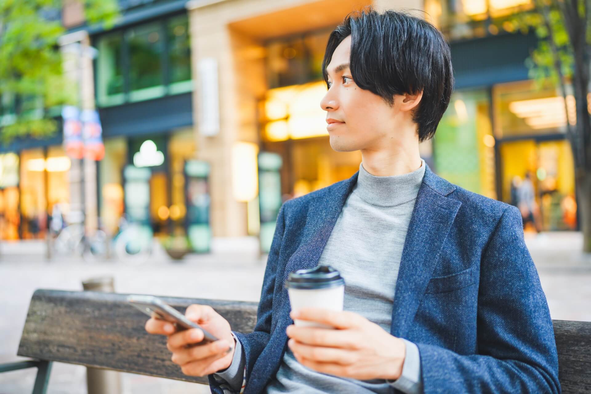 ネイビーのジャケットにタートルネックを合わせ、コーヒーを手に街で過ごす男性