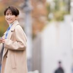 ベージュのロングコートを着て、スマートフォンを手に街で微笑む女性