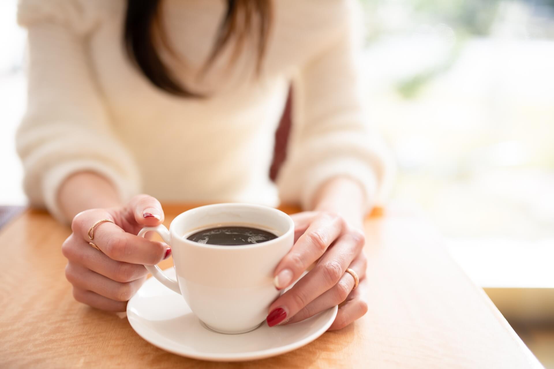 ニットを着た女性が、赤いネイルの手でコーヒーカップを持っている手元