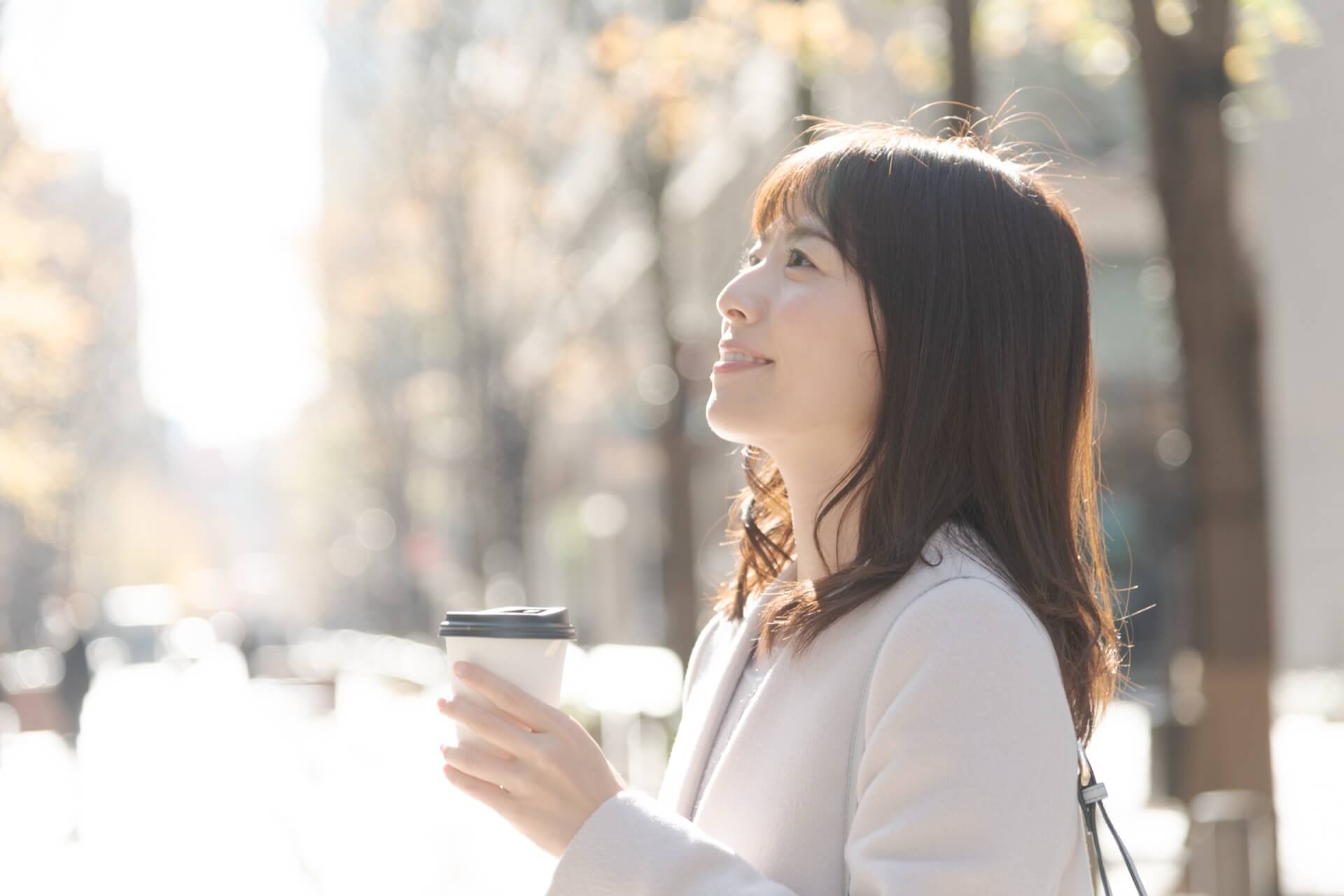 アイボリーのコートを着て、コーヒーを手に街を歩く女性